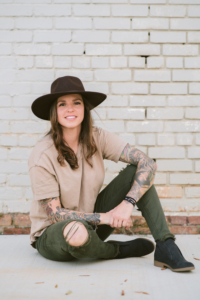 Girl with large brim hat sitting on the ground in front of a white brick wall wearing green ripped pants and a tan shirt with a full sleeve of tattoos.