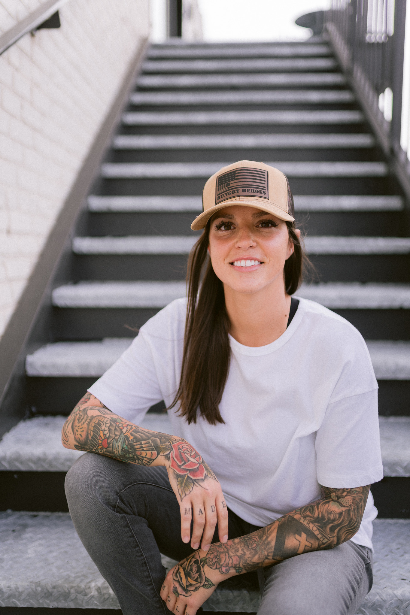 Girl with long brown hair wearing a tan hat sitting on concrete stairs with full sleeve tattoos smiling.