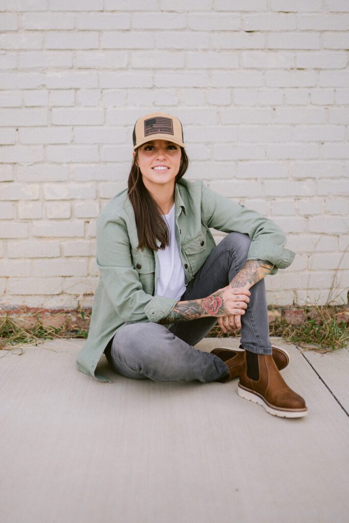 Woman sitting in front of a white brick wall with a tan hat and long sleeve sage green shirt and jeans with brown boots smiling